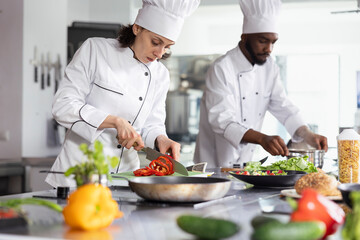 Woman chef in uniform slicing a bell pepper in professional kitchen, using fresh produce and kitchen tools to create a delicious dish. Culinary expert prepared ingredients on the stove.