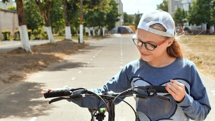 Obraz premium Smiling school girl wearing glasses and white cap holding bicycle handlebars, enjoying a sunny day outdoors