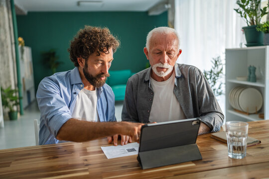 Adult son helping senior father with digital tablet and paperwork at home - Powered by Adobe