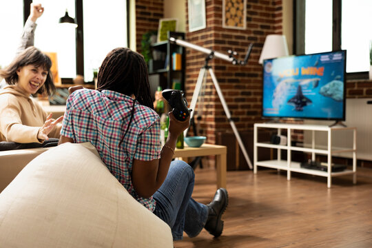 Excited young women celebrate shared win inside brick wall apartment during thrilling gaming console match. Cheerful friends seated on soft couch enjoying treats and expressing joy.