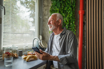 happy senior man using smart phone in kitchen, contemplating next move