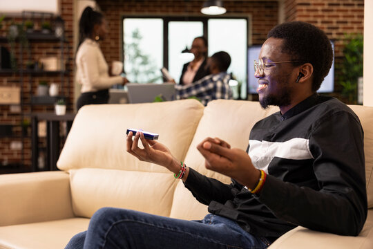Young black businessman sits on cozy sofa, projecting research data from handheld device while showcasing innovation in workplace. He wears glasses and earphones, engaging in futuristic presentation.