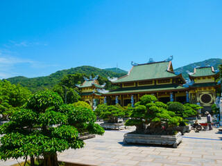 Obraz premium Traditional Vietnamese temple courtyard at Linh Ung Pagoda features upturned eaves, dragon motifs, vibrant roof tiles, bonsai trees in large stone planters, ornate architecture in Danang