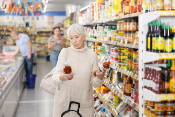 Attentive old woman purchaser choosing tomato juice out of large stock in supermarket