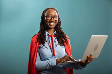 Portrait of black woman in blue shirt and red cape, standing with laptop in hand, representing a corporate superhero. African american female individual holds digital device and looks at camera.