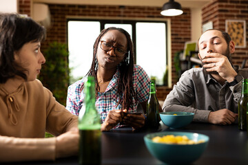 African american woman with curious expression looks at white female friend during casual game day. Multiethnic young adults enjoying indoor leisure time with card game, cold drinks and snacks.