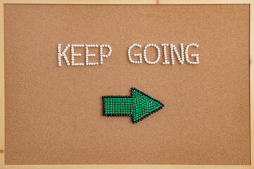 Inspirational message “Keep Going” spelled with push pins on a corkboard, featuring a green arrow pointing right. Symbol of motivation, direction, perseverance, and encouragement.