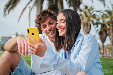 A cheerful couple is sitting together, enjoying a delightful moment while using a smartphone to...