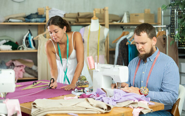 Professional tailor stitching custom clothing at sewing machine, while young female colleague skillfully cutting vibrant fabric on work table nearby in modern design studio....