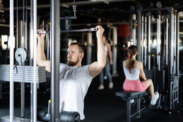 Fototapeta premium Athletic guy work out muscles of hands and arms on cable crossover machine in fitness club. Gym visitor trains tris, during classes at multi-station in wellness center.