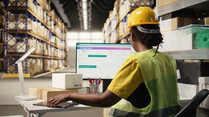 African american female working in logistics hub with tracking info on pc, updating the delivery route and other shipment details in real time. Following parcel status for online orders. Camera A.