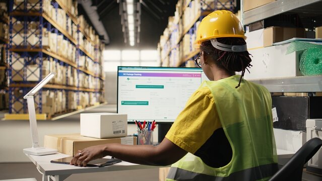African american female working in logistics hub with tracking info on pc, updating the delivery route and other shipment details in real time. Following parcel status for online orders. Camera A. - Powered by Adobe