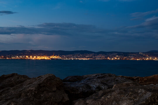 Panoramic view of the city and port of Trieste at dusk, with the lights reflecting on the sea. The photo was taken from Muggia, in Friuli-Venezia Giulia.