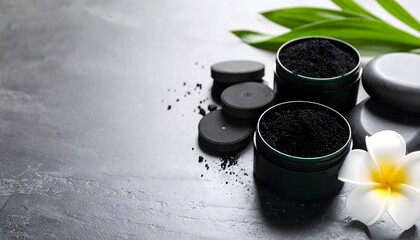 Artistic spa composition featu activated charcoal powder in containers alongside a frangipani flower on a textured gray background to evoke wellness and beauty.