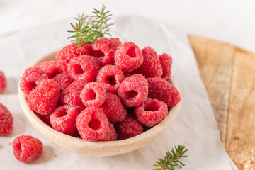 A rustic ceramic bowl filled with plump, fresh raspberries sits on a white table with a wooden board and scattered berries