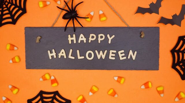 Happy Halloween sign with candy corn, decor and walking spider. Top view, stop motion video on an orange background.