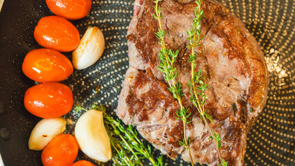 Close-up shot of a juicy ribeye steak sizzling in a frying pan with fresh thyme, garlic, and cherry...