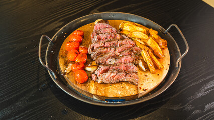Juicy sliced medium rare ribeye steak served on a wooden board with crispy homemade fries, roasted garlic, fresh thyme, and cherry tomatoes. Perfect for food blogs and menus.