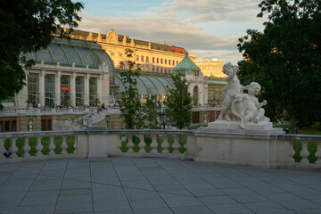 Fototapeta premium View of Vienna’s Palm House from a balustrade with infant sculptures in the foreground and a sunlit classical building in the background.