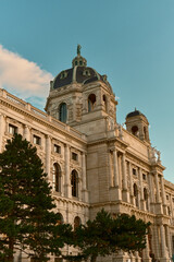 View of Vienna’s Kunsthistorisches Museum with pine trees in the foreground and a clear blue sky, highlighting the classical architecture and serene surroundings.