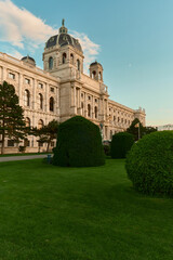 Obraz premium Scenic view of Vienna’s Kunsthistorisches Museum with lush green lawn in the foreground, surrounded by manicured shrubs and pine trees under a blue sky.