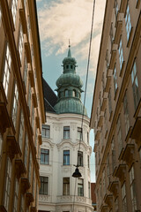 Obraz premium Upward view of a narrow Viennese street with classic facades, a black street lamp overhead, and a white building with a green dome and spire in the distance.
