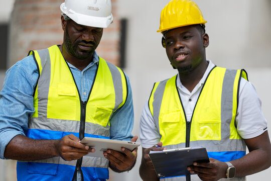 An experienced construction foreman mentors a young apprentice on a building site. The senior engineer uses a tablet to explain project details, providing on-the-job training and guidance.