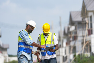 An experienced construction foreman mentors a young apprentice on a building site. The senior engineer uses a tablet to explain project details, providing on-the-job training and guidance.
