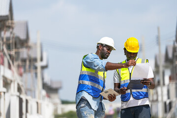 An experienced construction foreman mentors a young apprentice on a building site. The senior engineer uses a tablet to explain project details, providing on-the-job training and guidance.