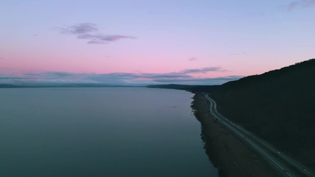 Pink night sky over the lake with highway and cars with headlights on passing below. Wind turbines line the lefthand side of the lake. 