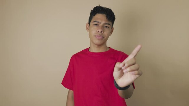 Young Latino man moving his index finger side to side in a gesture of denial on a light brown background. Concept of rejection, disapproval, warning, and personal boundary