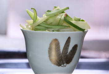 A Detailed Close-up, Eye-Level Shot of an Elegant White Bowl with Gold Feather Patterns, Filled with a Fresh and Appetizing Pile of Sliced Green and White Vegetables or Fruit