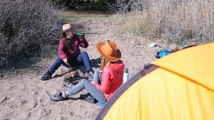 Two women are sitting on the beach next to a tent. Two alcoholics are having fun on the lake shore. Sisters roast sausages at a picnic