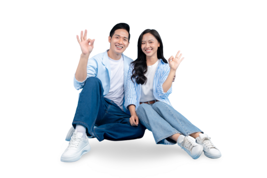 A happy couple is sitting close together and gesturing OK signs on PNG background. They are smiling and wearing light blue clothing. - Powered by Adobe