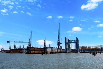 Obraz premium A striking view of a shipyard featuring towering cranes, strong structures, and glistening water, all under a bright blue sky with scattered clouds. Perfect for industrial project inspiration.