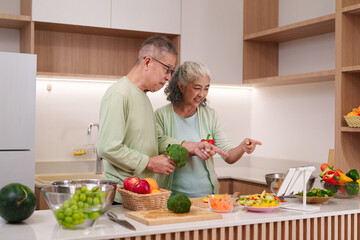 Asian senior couple preparing healthy meal in modern kitchen using tablet for recipe guidance older adults cooking fresh vegetables fruit at home