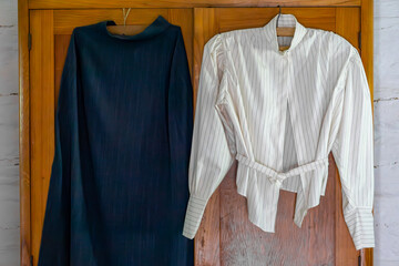 Traditional clothing hanging on a wooden wall – a dark long robe and a white striped shirt on...