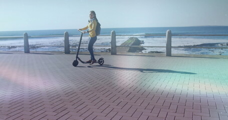 Riding female wearing yellow jacket on e-scooter along seaside promenade, with backpack and scarf