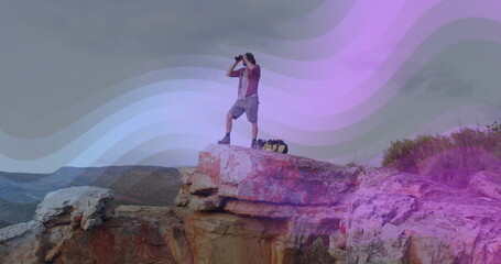 Scanning lone hiker wearing boots using binoculars while standing on sandstone cliff, with backpack