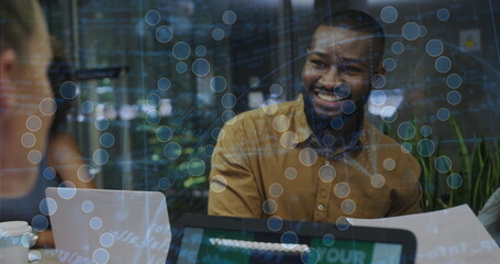 Smiling man in mustard top talking at office table with laptops, papers, mug, glass network overlay