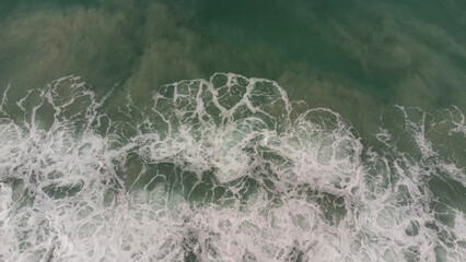 Aerial view of ocean waves crashing with white foam on green sea surface, creating natural texture and abstract pattern, perfect for backgrounds, nature concepts, and coastal design.