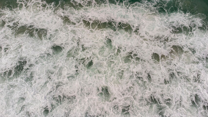 Aerial view of ocean waves crashing with white foam on green sea surface, creating natural texture and abstract pattern, perfect for backgrounds, nature concepts, and coastal design.