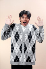 Asian man in gray argyle cardigan raising both hands with surprised facial expression, open mouth,...