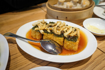 Steamed tofu topped with mushrooms and greens in savory soy-based sauce, served on a white plate...