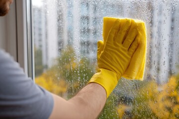 A person wearing yellow gloves cleans a window with a yellow cloth droplets visible