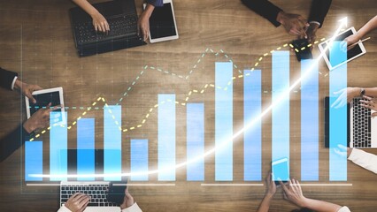 A business team engages in collaborative performance analysis using digital devices while reviewing growth graphs and statistics on a wooden desk in an office setting. Copula
