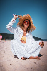 Stylish woman in straw hat, red sunglasses, and white summer outfit sitting on sandy beach, smiling under bright sun. Concept of summer fashion, holiday vibes, travel lifestyle, and joy.