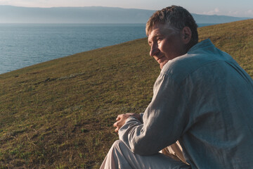 candid portrait of a man enjoys moment tranquility while seated on grassy slope. he gazes thoughtfully across serene lake, basking in warm light setting sun. close up.