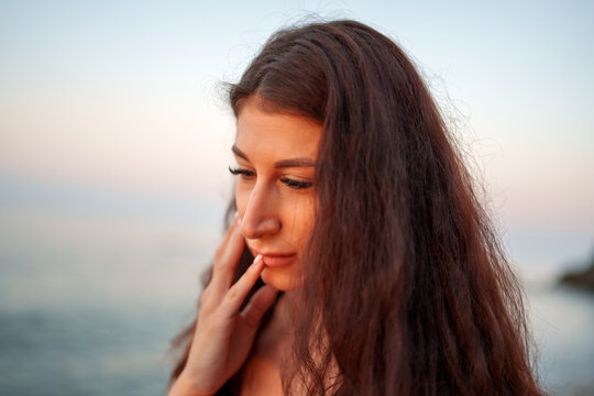 Sunset lit woman with flowing brown hair gently caressing cheek while gazing downward on sandy shoreline