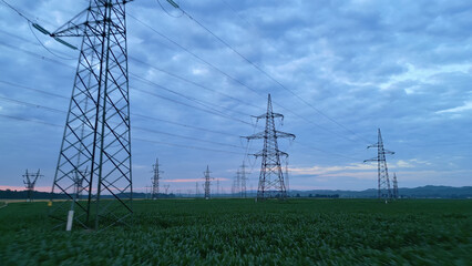 Establishing shot of electricity pylons across a green field under twilight, symbolizing sustainable energy infrastructure. Highlights nature and technology, modern power distribution.